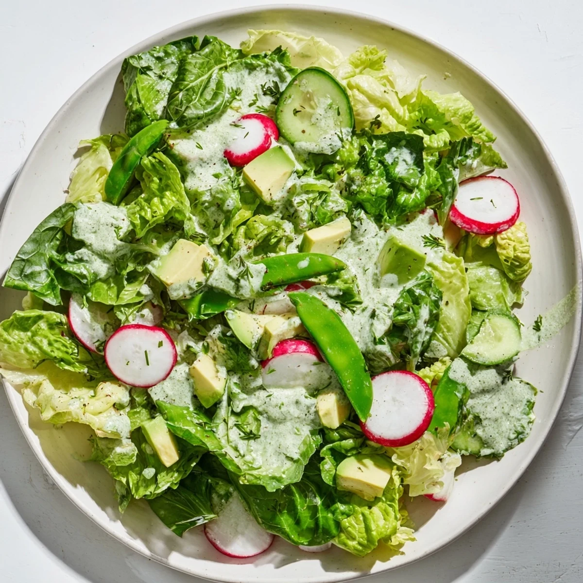 A vibrant Green Salad with Green Goddess Dressing featuring crisp greens, cucumber slices, and creamy avocado chunks, all generously tossed in a vibrant green herb dressing in a ceramic bowl.