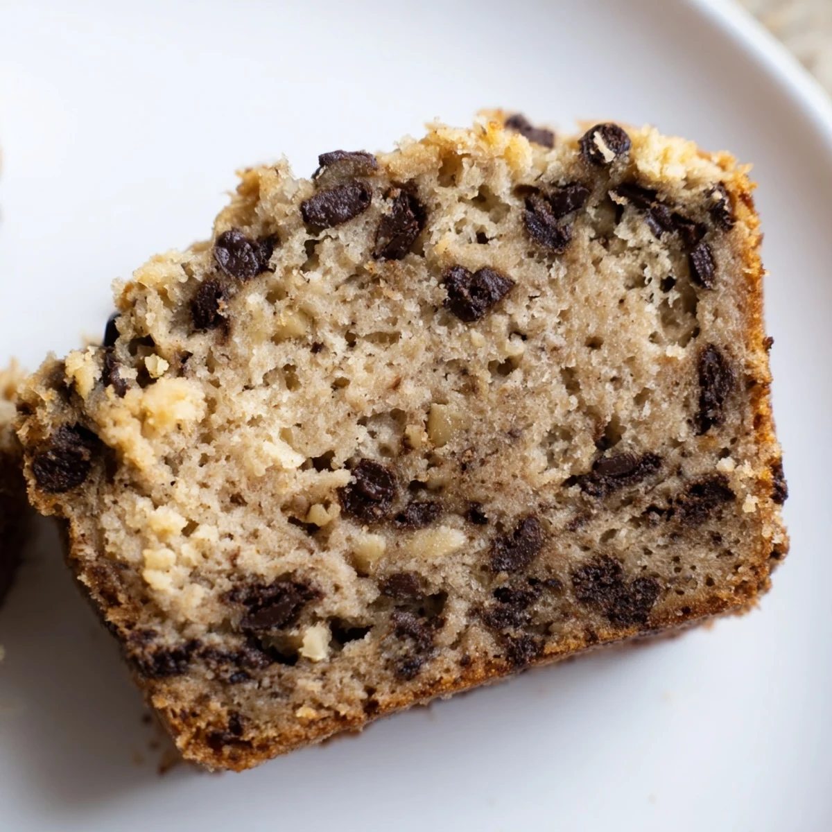 A slice of warm chocolate chip banana bread is slathered with melting butter, ready to enjoy with a steaming cup of coffee.