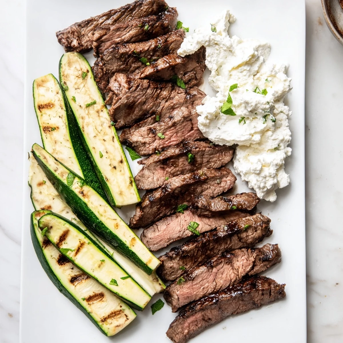 Balsamic Flank Steak With Grilled Zucchini And Whipped Feta plated alongside charred vegetables and herbs for a Mediterranean dinner.