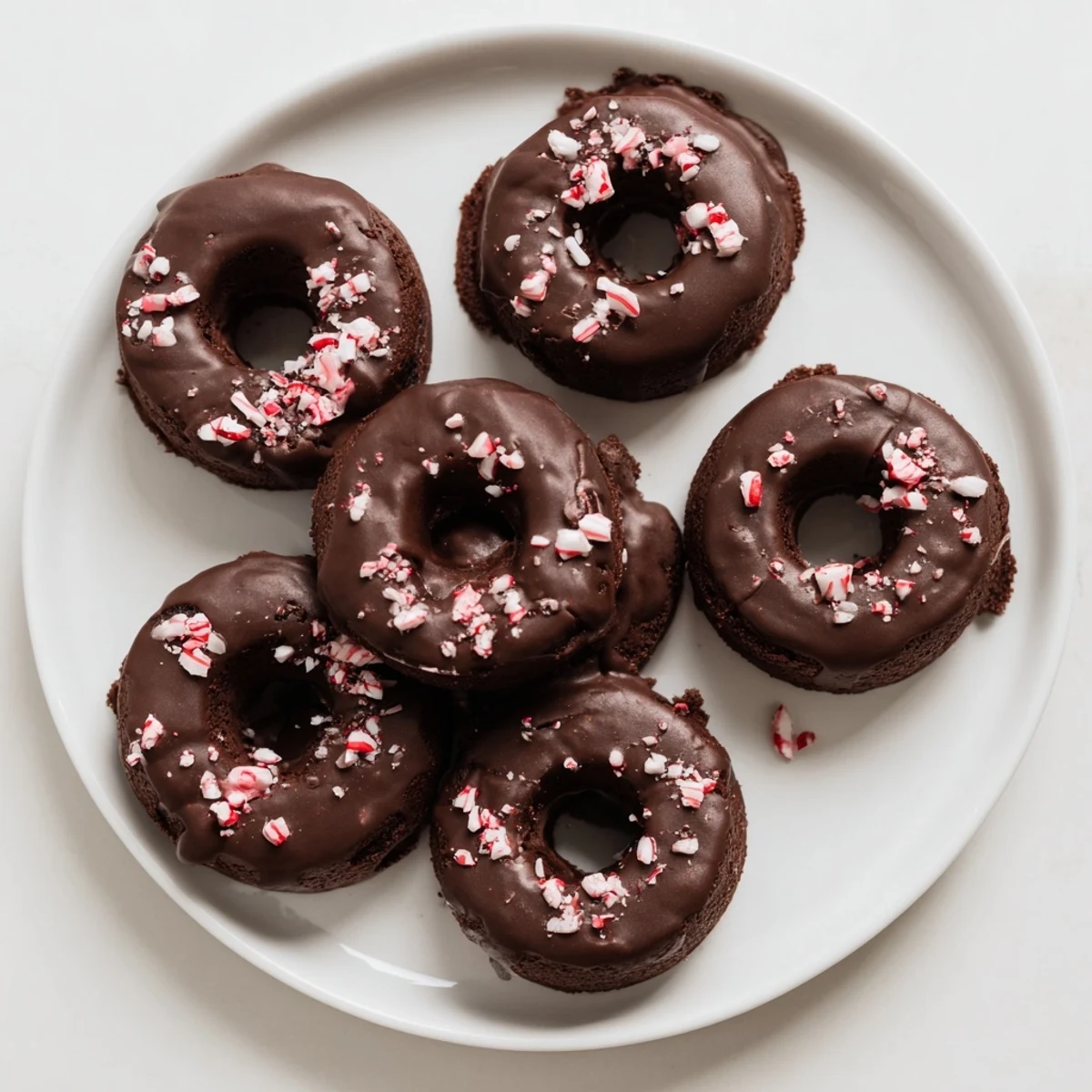Four freshly baked Chocolate Peppermint Mochi Donuts with a glossy glaze and crushed candy canes sit on a cooling rack.