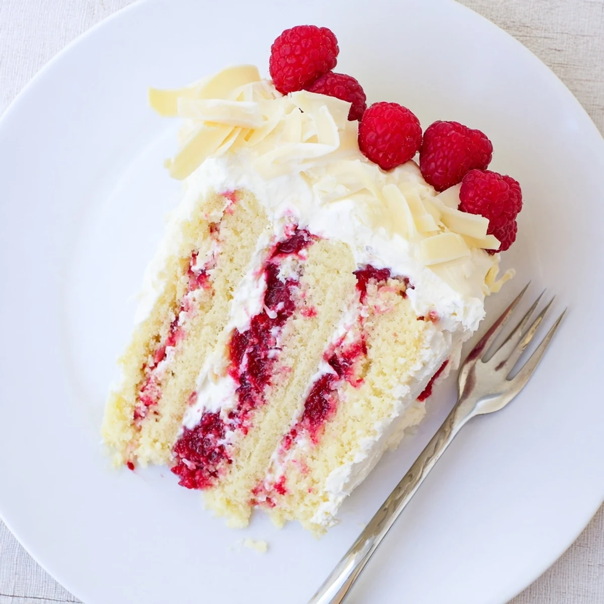 Decadent white chocolate raspberry cream cake with whipped cream icing and fresh raspberries garnished on a white serving plate