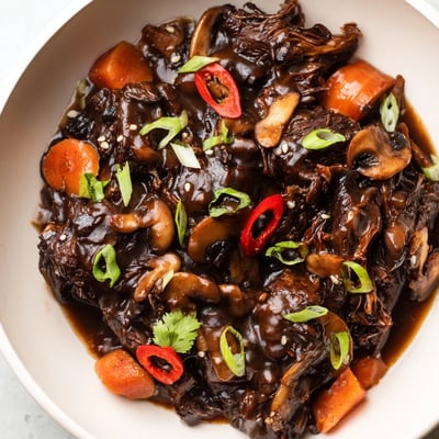 Steaming Asian Style Slow Cooked Beef Cheeks With Mushrooms feature fork-tender braised beef coated in a savory umami sauce, with chunks of carrots and cremini mushrooms alongside a garnish of fresh green onions and cilantro.