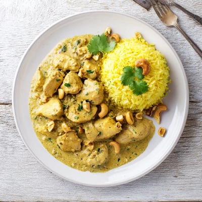 A close-up of tender chicken simmered in a rich coconut cashew sauce next to fluffy, saffron-infused rice and a cinnamon stick.